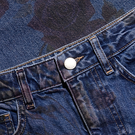 Close-up of the Mallis Bow-Leg Jean in blue denim, featuring a metallic button and faint rose floral patterns on the fabric.