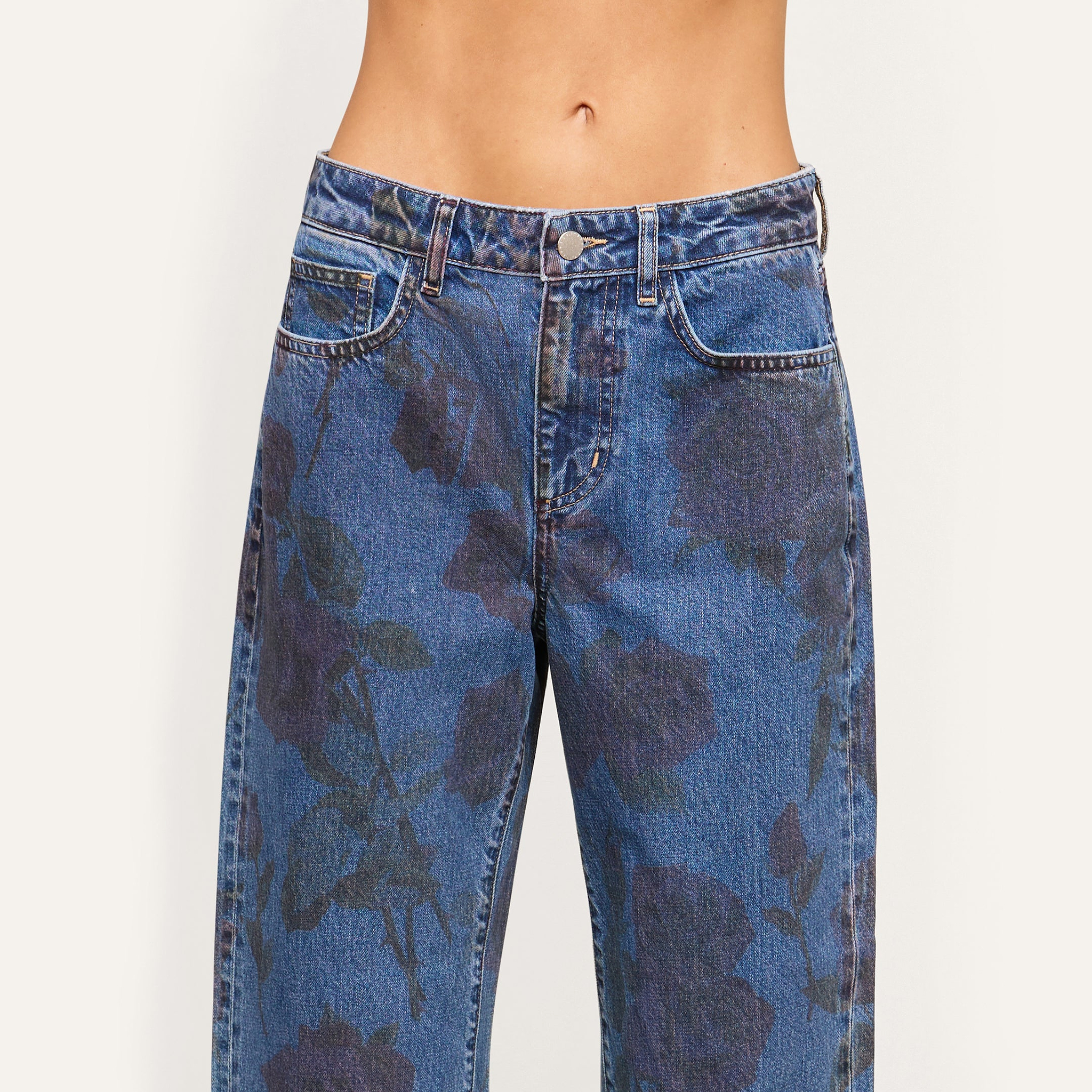 A person wearing the Mallis Bow-Leg Jean, a high-waisted blue jean featuring a subtle dark rose print and a wide-leg silhouette; their torso is visible from the waist to just above the navel.