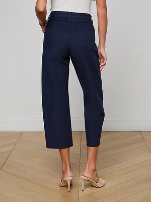 A person wearing the Mallis Bow-Leg Jean in a dark indigo wash and beige high-heeled shoes stands on a wooden floor, shown from the waist down and facing away from the camera.