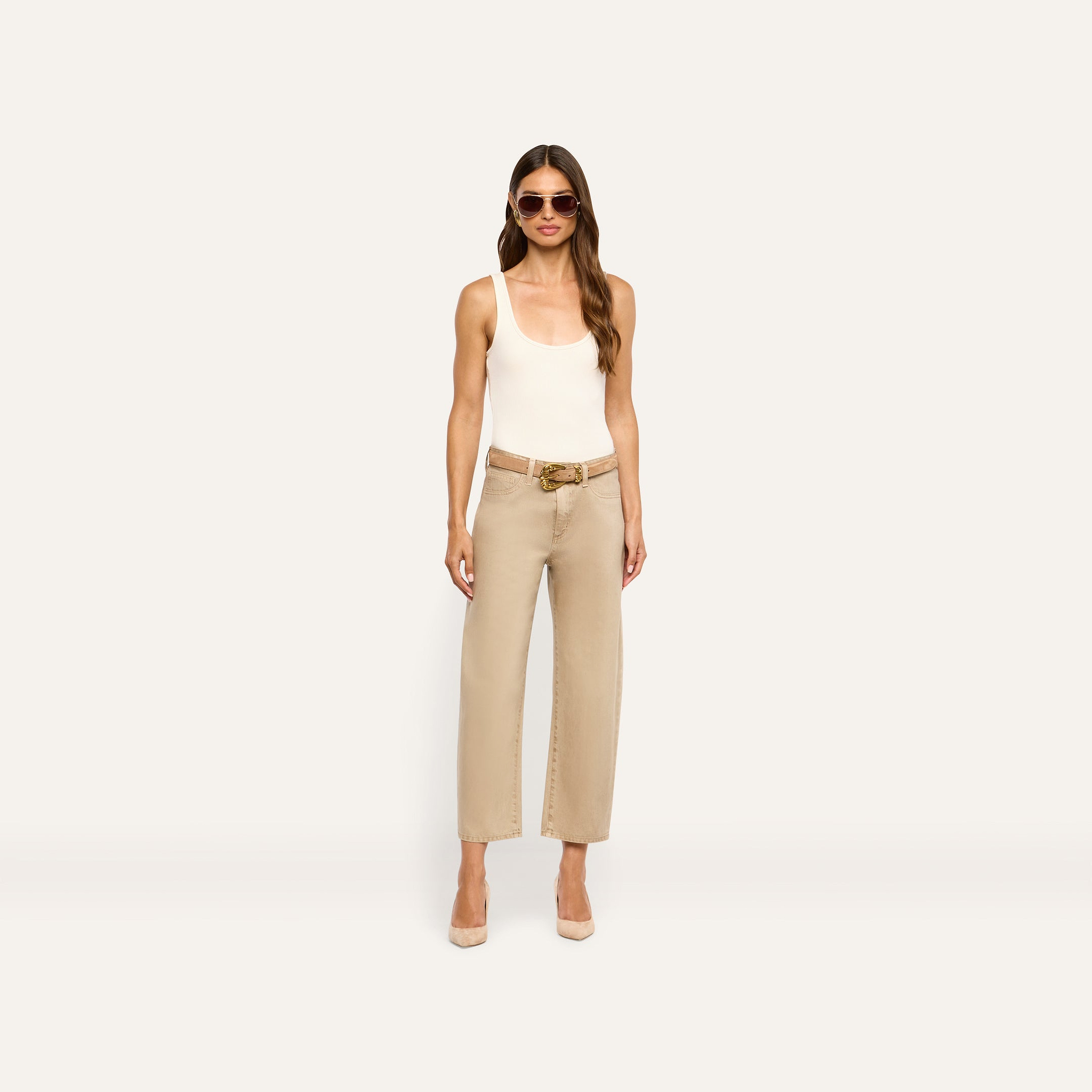 A woman wearing a white tank top, a tan belt with a gold buckle, and tan Mallis Coated Bow-Leg Jean with heels.
