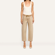 A person wears a beige tank top and high-rise, wide-leg tan Mallis Coated Bow-Leg Jean with a gold-buckled belt and tan pointed-toe heels.
