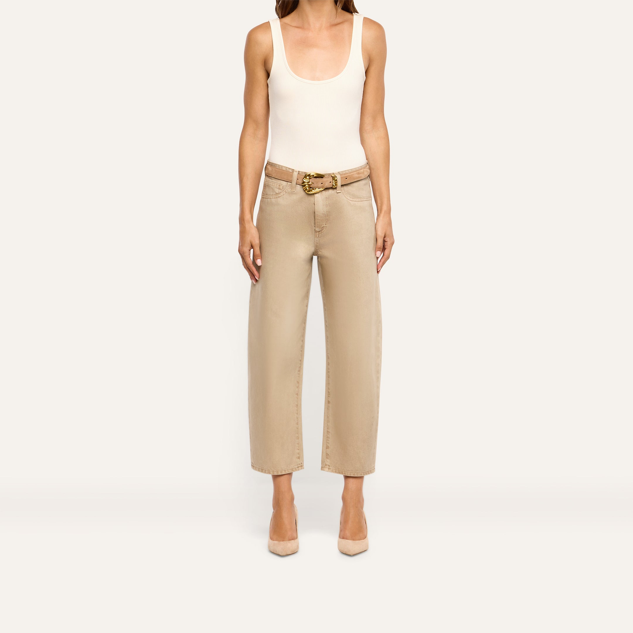 A person wears a beige tank top and high-rise, wide-leg tan Mallis Coated Bow-Leg Jean with a gold-buckled belt and tan pointed-toe heels.