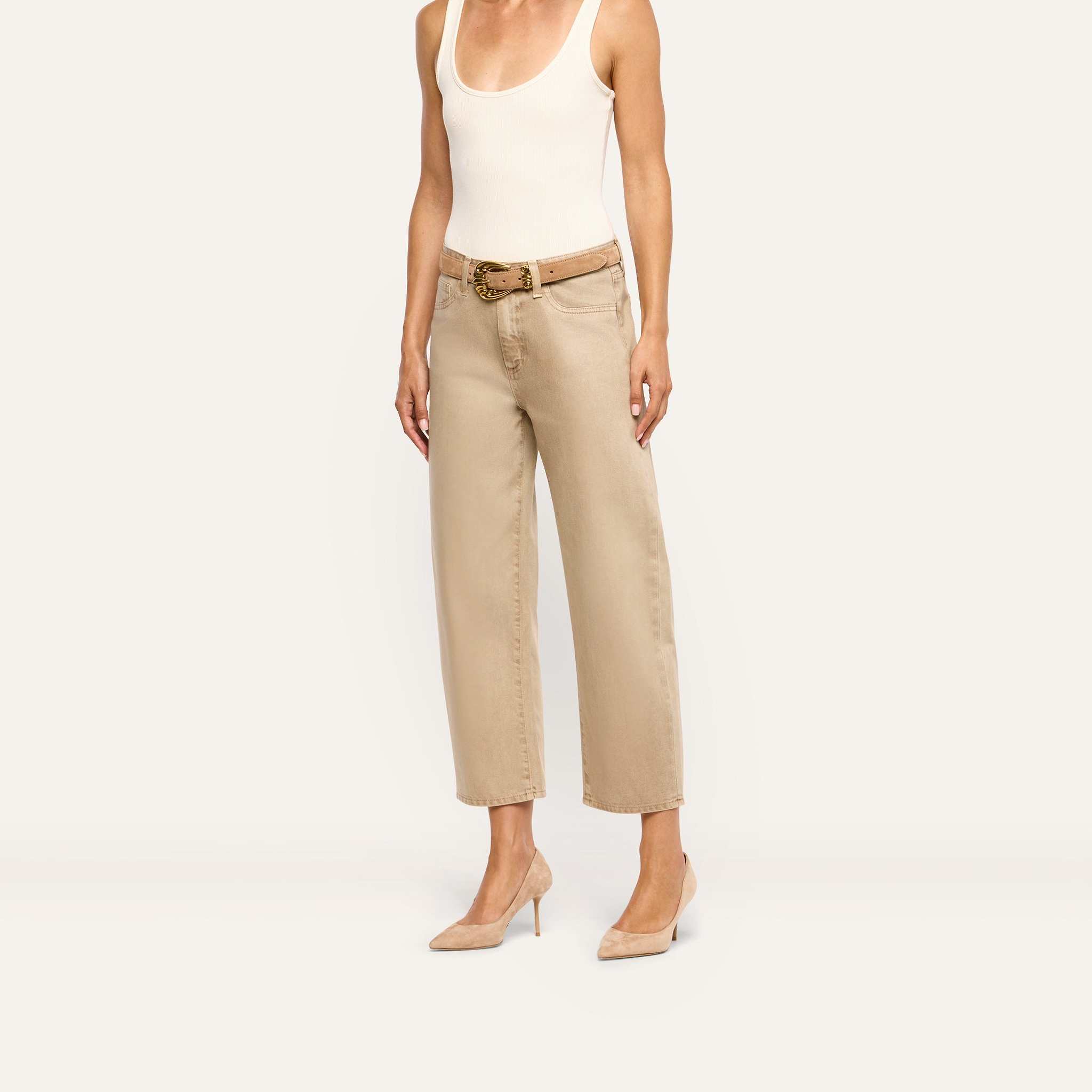 A person wears a white tank top, a tan belt with a large buckle, Mallis Coated Bow-Leg Jean, and tan pointed-toe heels.