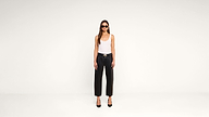 A woman wearing sunglasses, a white tank top, black leather pants, and black heels stands in a minimal, white studio setting, facing forward with arms relaxed at her sides.