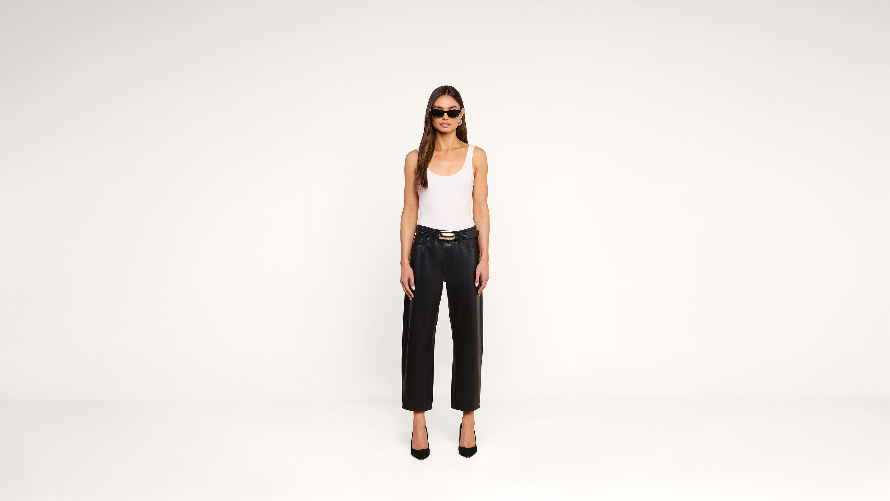 A woman wearing sunglasses, a white tank top, black leather pants, and black heels stands in a minimal, white studio setting, facing forward with arms relaxed at her sides.
