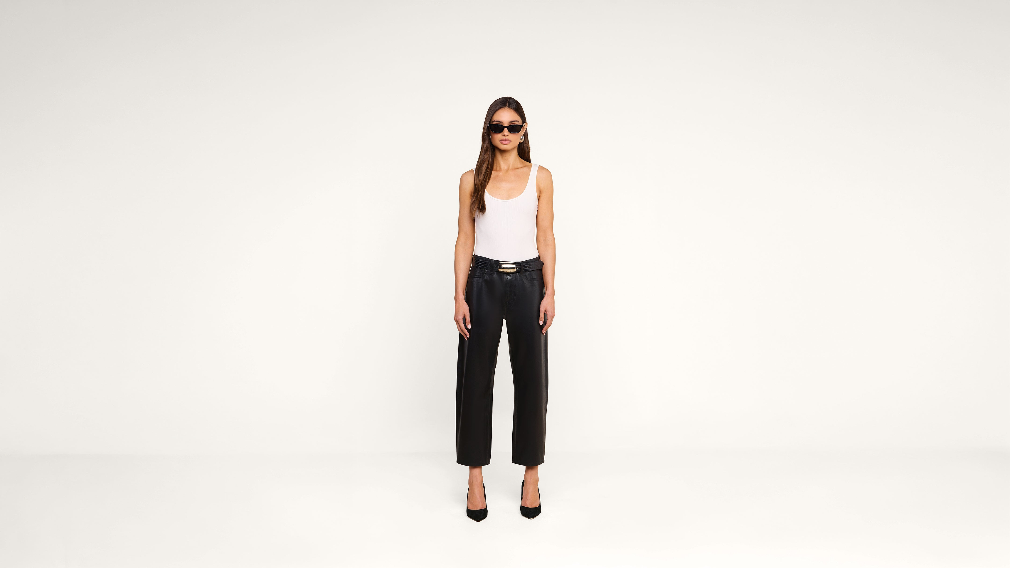 A woman wearing sunglasses, a white tank top, black leather pants, and black heels stands in a minimal, white studio setting, facing forward with arms relaxed at her sides.