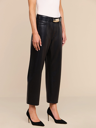 Wearing the Mallis Coated Bow-Leg Jean, a person stands against a beige background styled with a black belt featuring a gold buckle, a white sleeveless top, and black high heels.