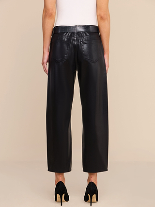A person stands against a beige background, wearing the Mallis Coated Bow-Leg Jean—a high-rise black coated denim pant with wide legs—paired with black heeled shoes. The photo is taken from the back, highlighting the fit and pocket details of the jeans.