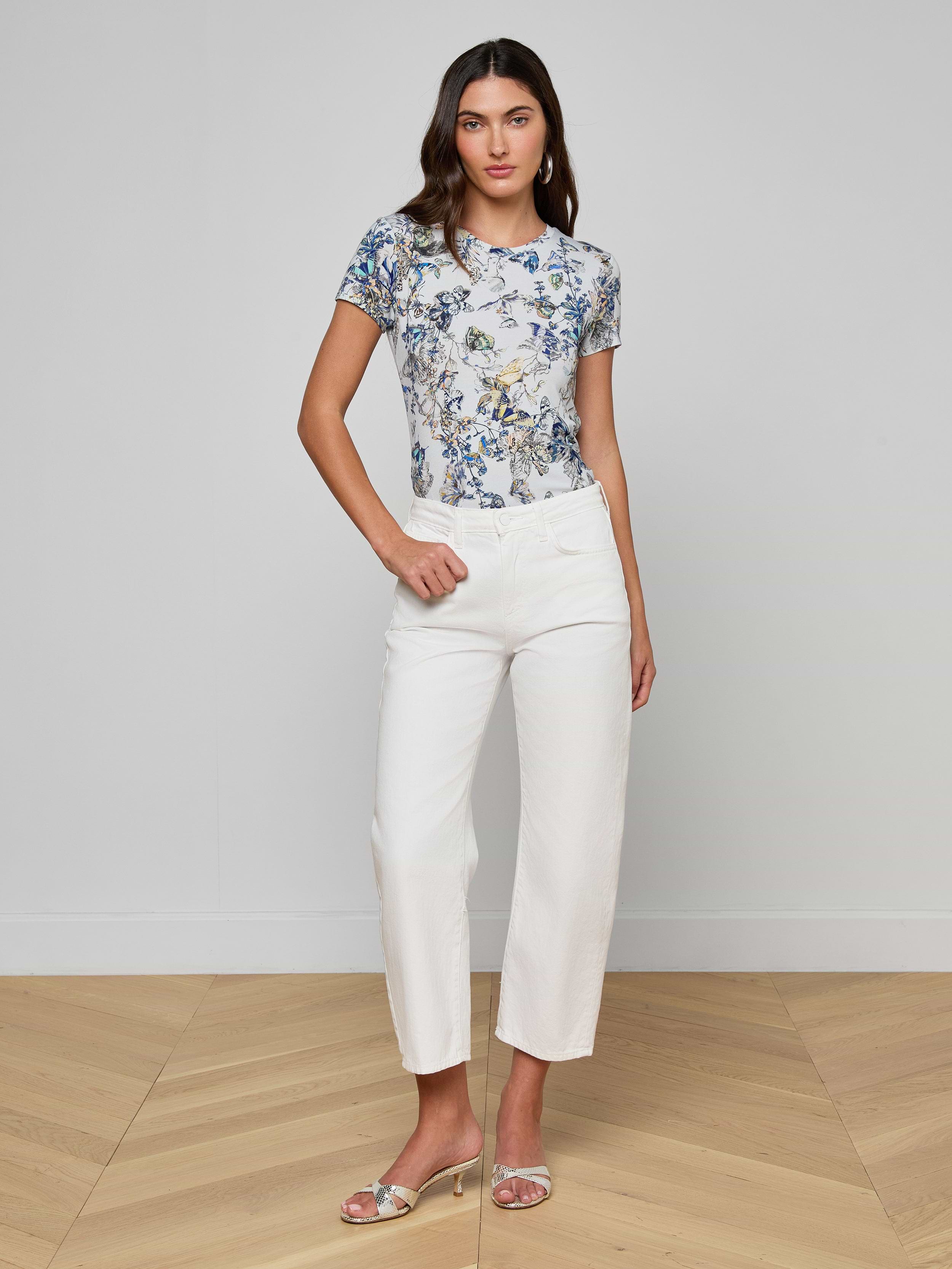 A woman stands against a plain white background, wearing a short-sleeve blue floral top, the Mallis Bow-Leg Jean in a high-rise wide-leg denim style, and open-toe heels. She has one hand on her hip and a neutral expression.