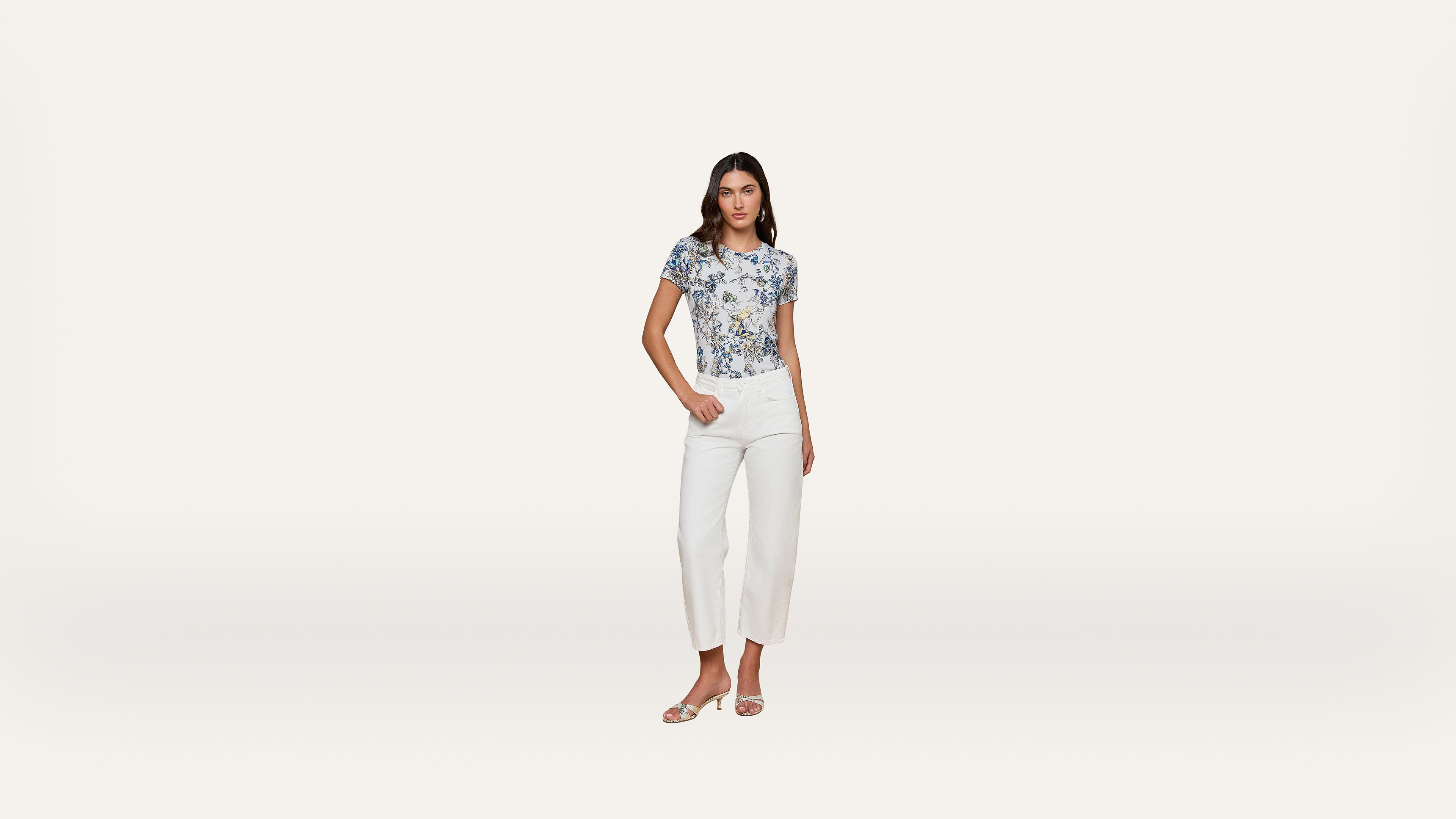 A woman wearing a floral top, white pants, and sandals stands facing forward against a plain white background.