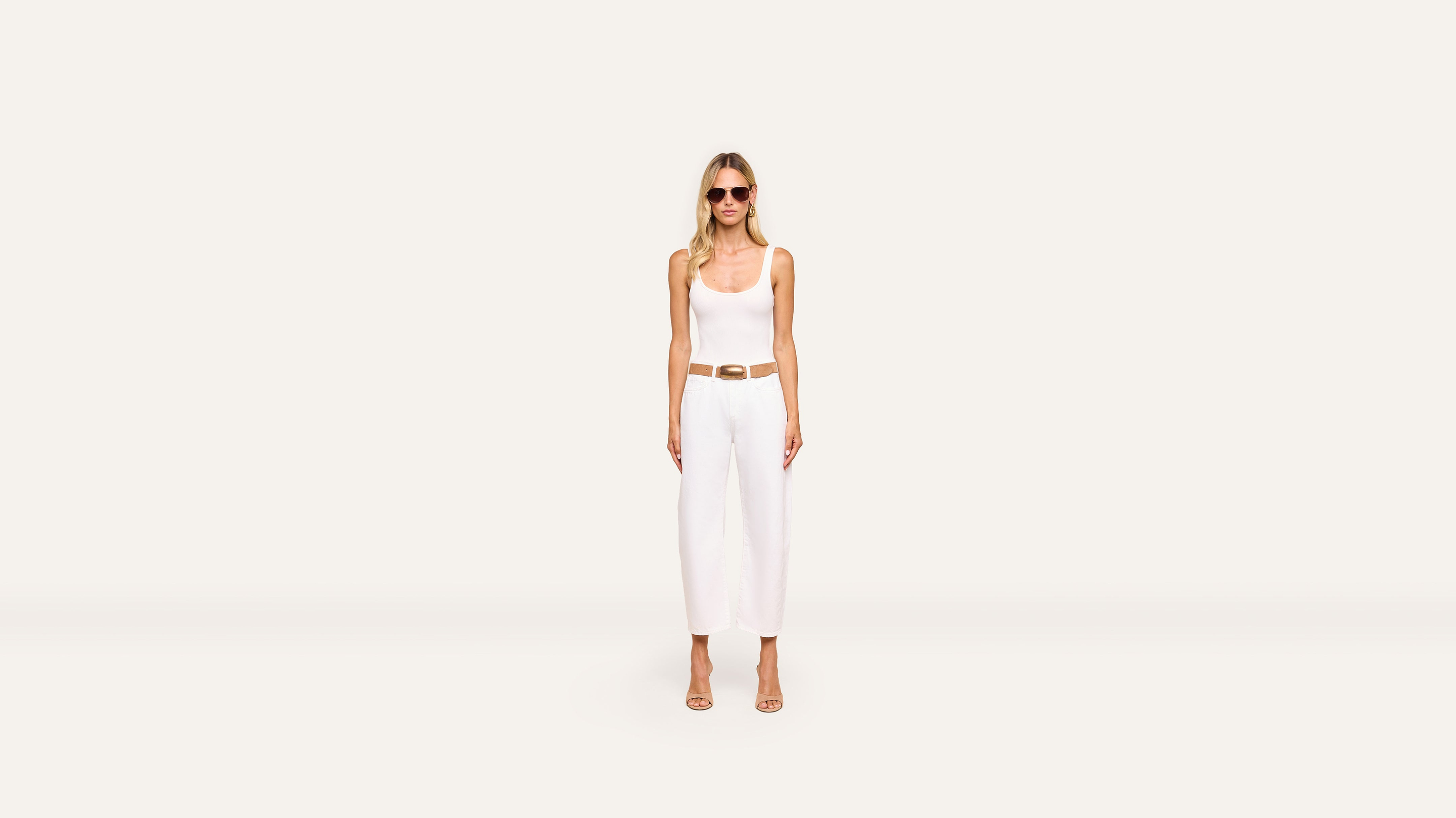 A woman in sunglasses, a white sleeveless top, white wide-leg pants, and beige heels stands against a plain, light background, accentuating her look with a brown belt.