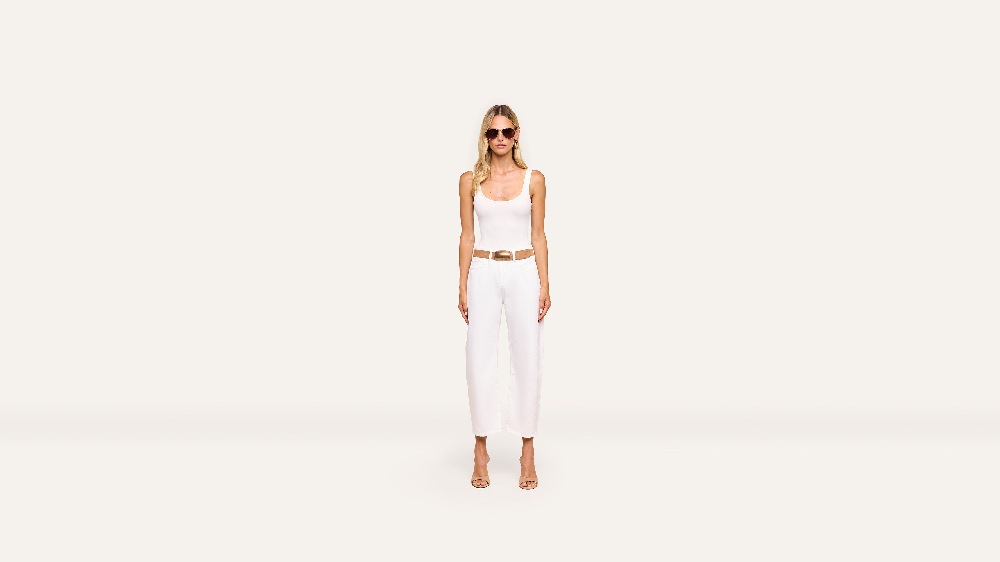 A woman in sunglasses, a white sleeveless top, white wide-leg pants, and beige heels stands against a plain, light background, accentuating her look with a brown belt.