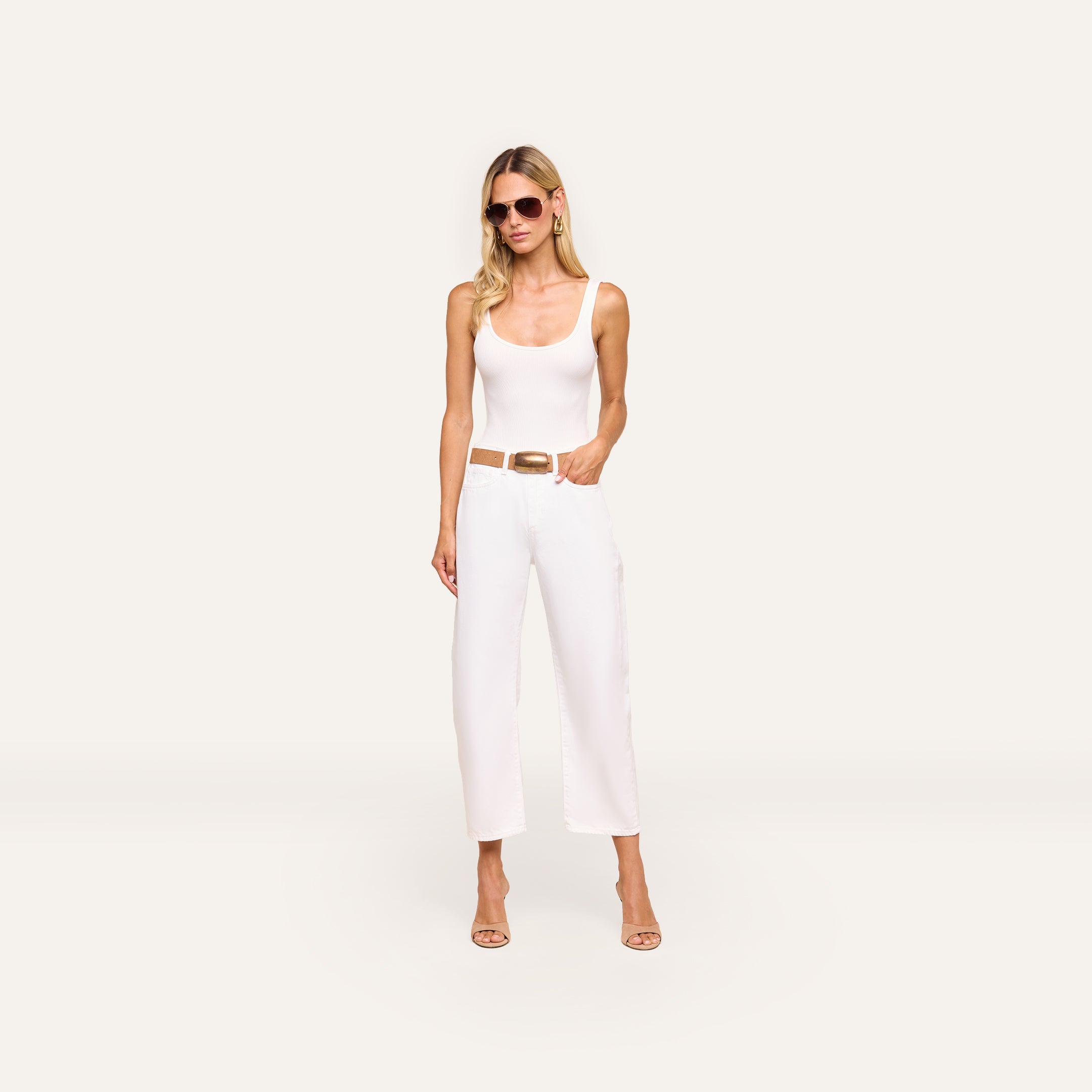 Woman wearing sunglasses, a white tank top, and Mallis Bow-Leg Jean with a brown belt and beige heels stands against a plain light background with one hand in her pocket.