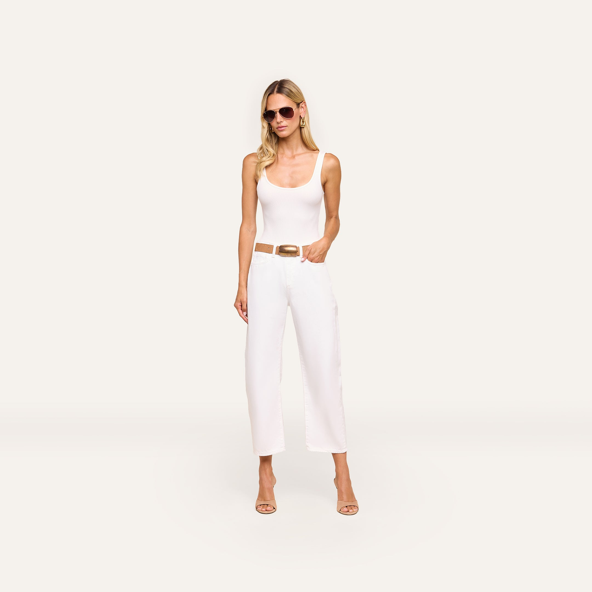 Woman wearing sunglasses, a white tank top, and Mallis Bow-Leg Jean with a brown belt and beige heels stands against a plain light background with one hand in her pocket.