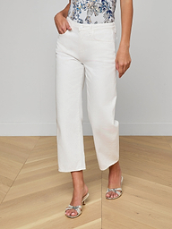 Wearing the Mallis Bow-Leg Jean in a high rise, white denim cropped style, paired with a patterned blue and white top and silver heeled sandals, a model’s lower body is shown standing against a plain white background.