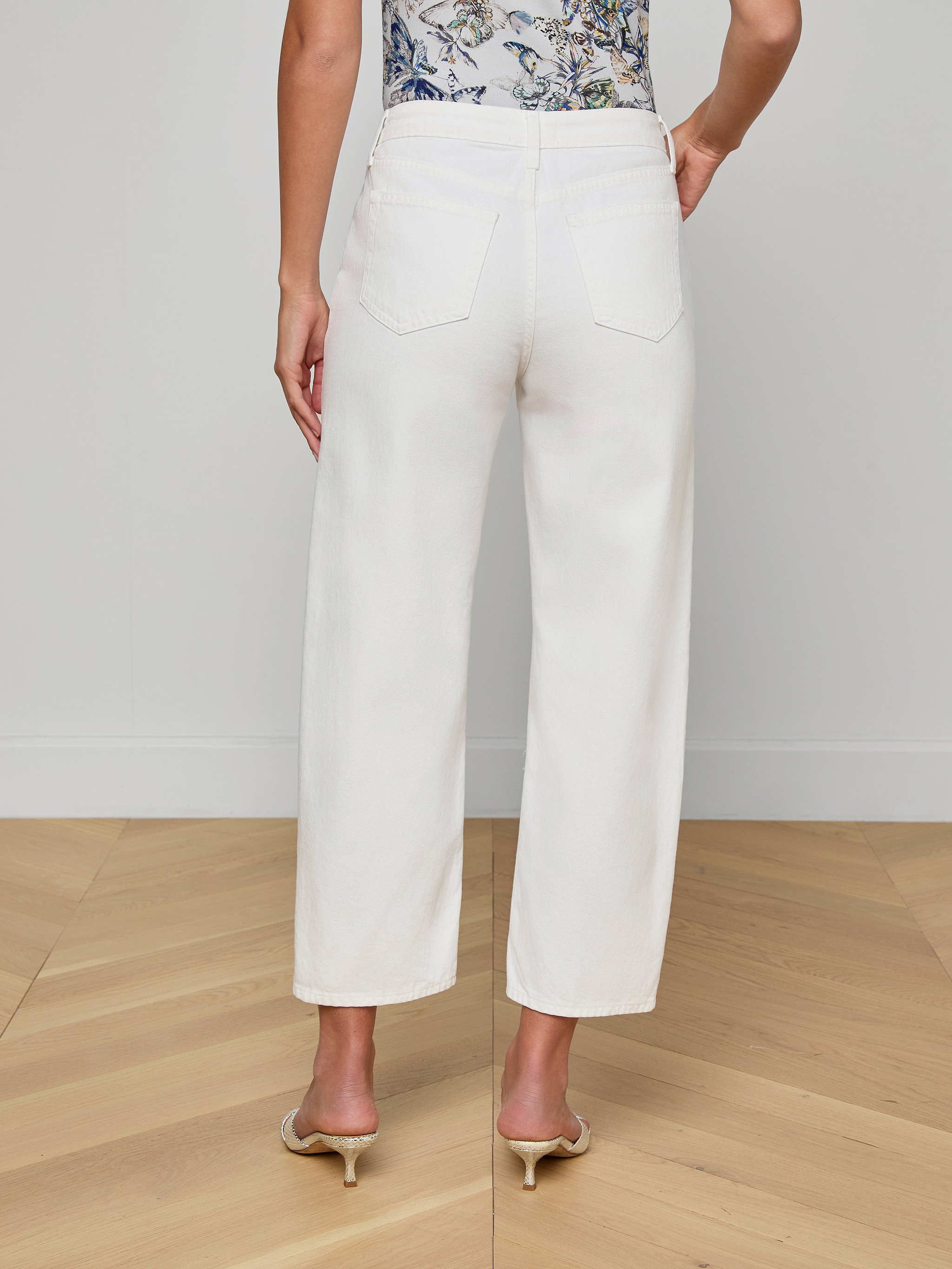 A person wearing the Mallis Bow-Leg Jean in white and beige heels stands with their back to the camera. The top part of a patterned shirt is visible, set against a plain, light-colored background.
