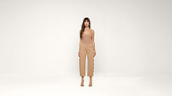 A woman stands in a neutral studio wearing a sleeveless nude top, high-waisted tan pants with side buttons, and nude heels. She faces forward with arms relaxed by her sides.