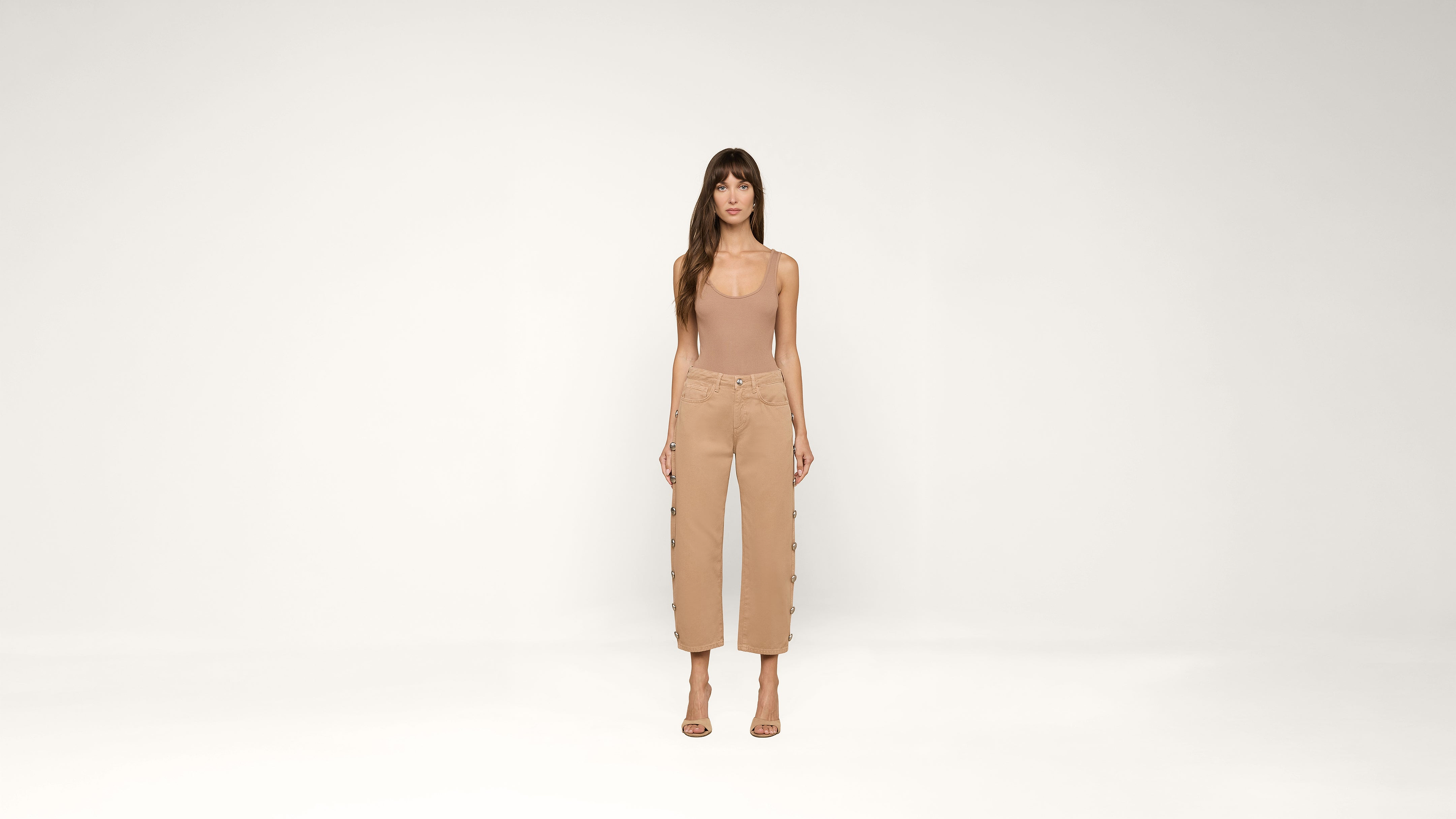 A woman stands in a neutral studio wearing a sleeveless nude top, high-waisted tan pants with side buttons, and nude heels. She faces forward with arms relaxed by her sides.