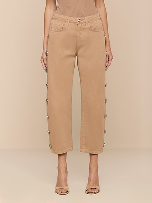 Wearing the Mallis Studded Bow-Leg Jean, featuring silver snap buttons along the side seams, a sleeveless beige top, and open-toed heels, a person stands against a neutral background.