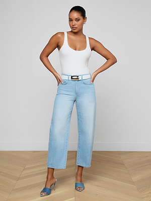 A woman stands confidently with her hands on her hips, wearing a white tank top, blue heeled sandals, and the Mallis Bow-Leg Jean in a light wash high rise wide-leg style, finished with a white belt against a plain background.