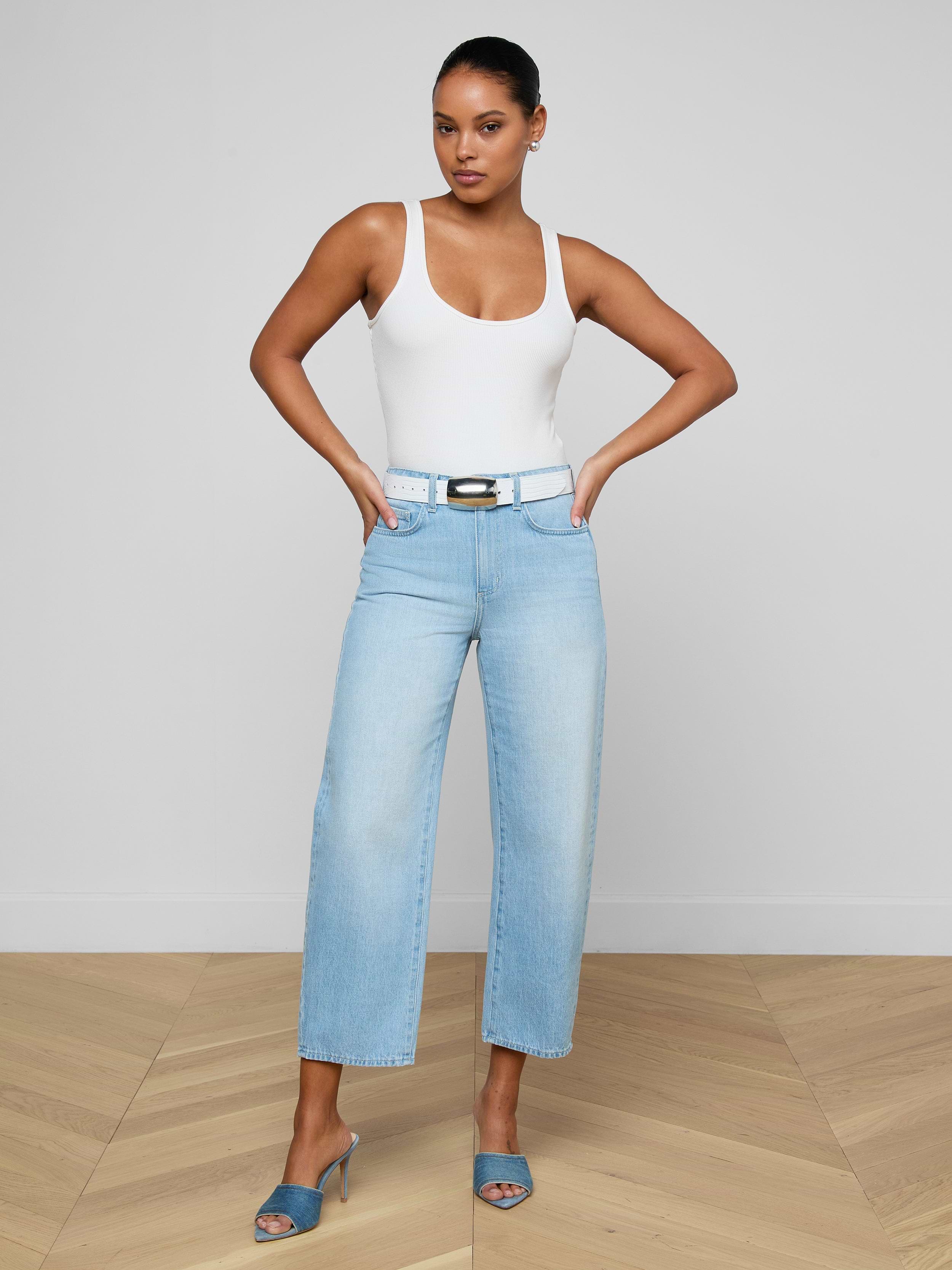 A woman in a white tank top, Mallis Bow-Leg Jean in light blue with a belt, and blue heeled sandals stands with hands on hips against a plain light background.