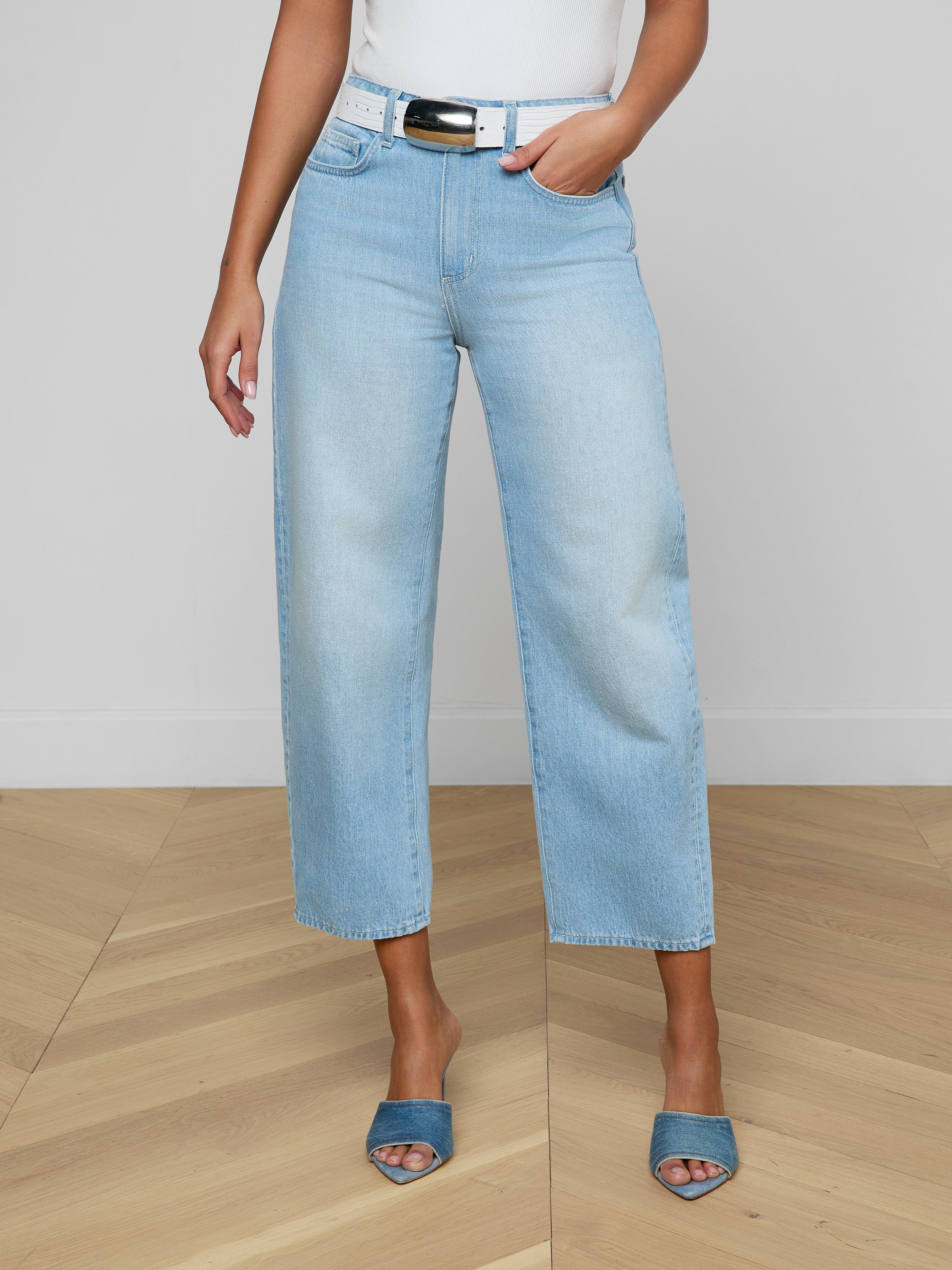 A person stands against a plain white background wearing the Mallis Bow-Leg Jean in light blue with a wide-leg, modern silhouette, paired with a white top and blue open-toe heels. The high-rise denim is styled fashionably, and the image is cropped above the shoulders.