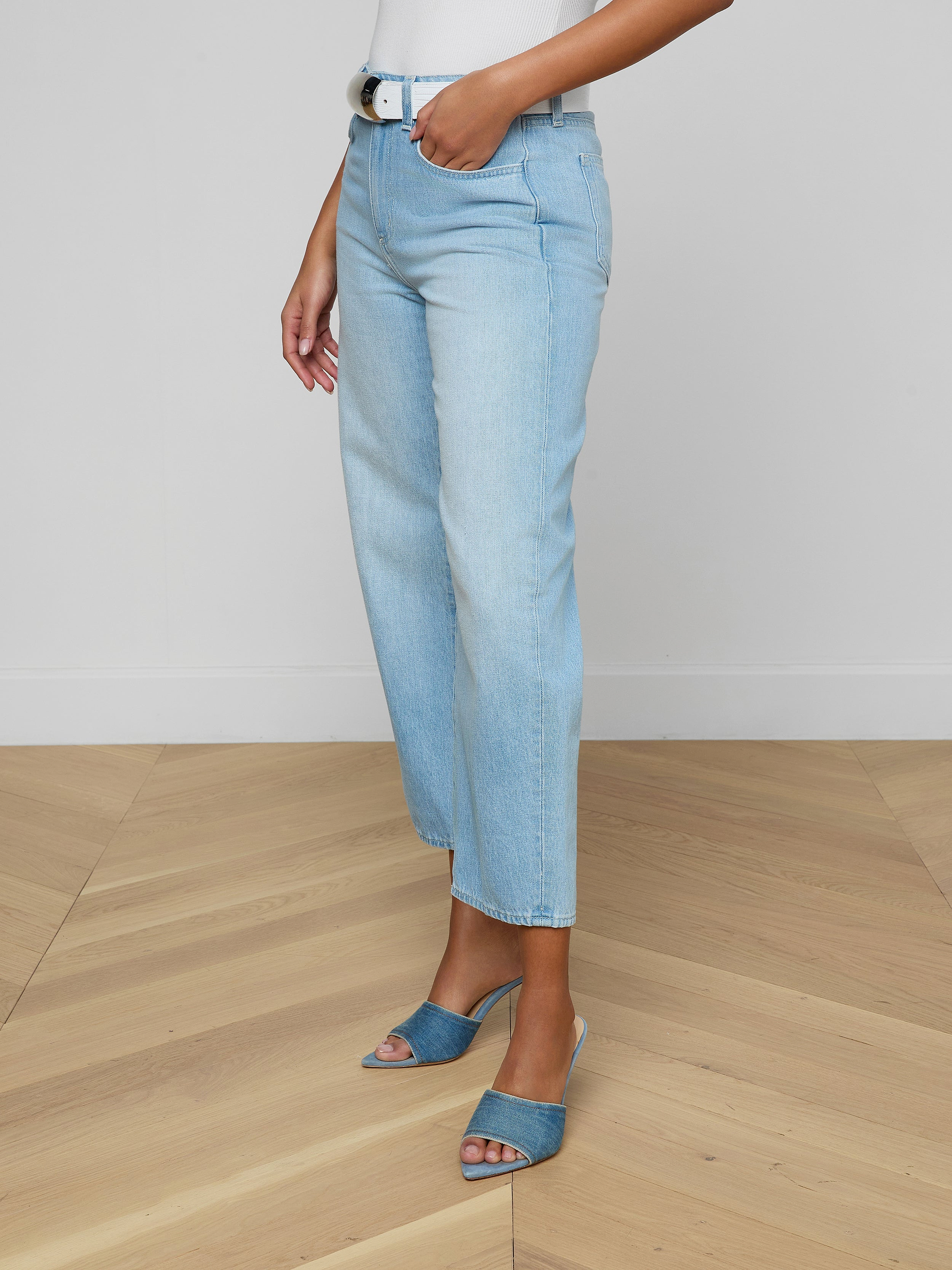 Woman wearing the Mallis Bow-Leg Jean in a light wash with a high rise and straight-leg fit, paired with a white top and blue open-toe heeled sandals, standing against a plain white background. Only the lower half of her body is visible.