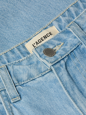 Close-up of the Mallis Bow-Leg Jean in light blue denim, featuring a high-rise waistband, silver button, and a white “L’AGENCE” tag. Light wash fabric texture is clearly visible.