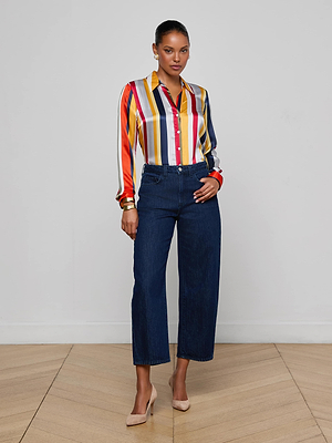 A woman stands indoors on a wooden floor, wearing the Mallis Bow-Leg Jean in dark blue, a colorful striped blouse, beige heels, and her hair pulled back with one hand on her hip.