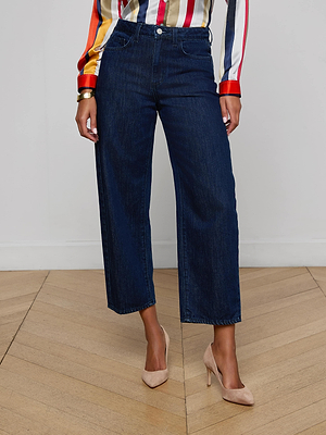 Someone is wearing the Mallis Bow-Leg Jean in dark blue, paired with a colorful striped long-sleeve blouse and beige high heels, standing on a light wooden floor against a plain white wall.