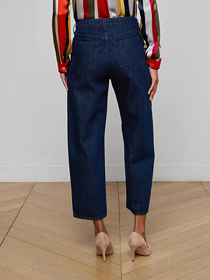 A person wearing Mallis Bow-Leg Jean in dark blue, a colorful striped long-sleeve shirt, and beige high-heeled shoes stands on a wooden floor facing away from the camera.