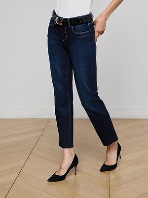 A person wearing Aiden Low-Rise Slim-Leg Jeans with a black belt, white top, and black high-heeled shoes stands on a wooden floor against a plain white wall.