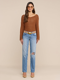 A woman wearing sunglasses, a brown long-sleeve top, and the Armida Low-Rise Slouch Jean—straight leg, ripped blue denim—paired with a tan belt and beige heels stands against a plain white background.