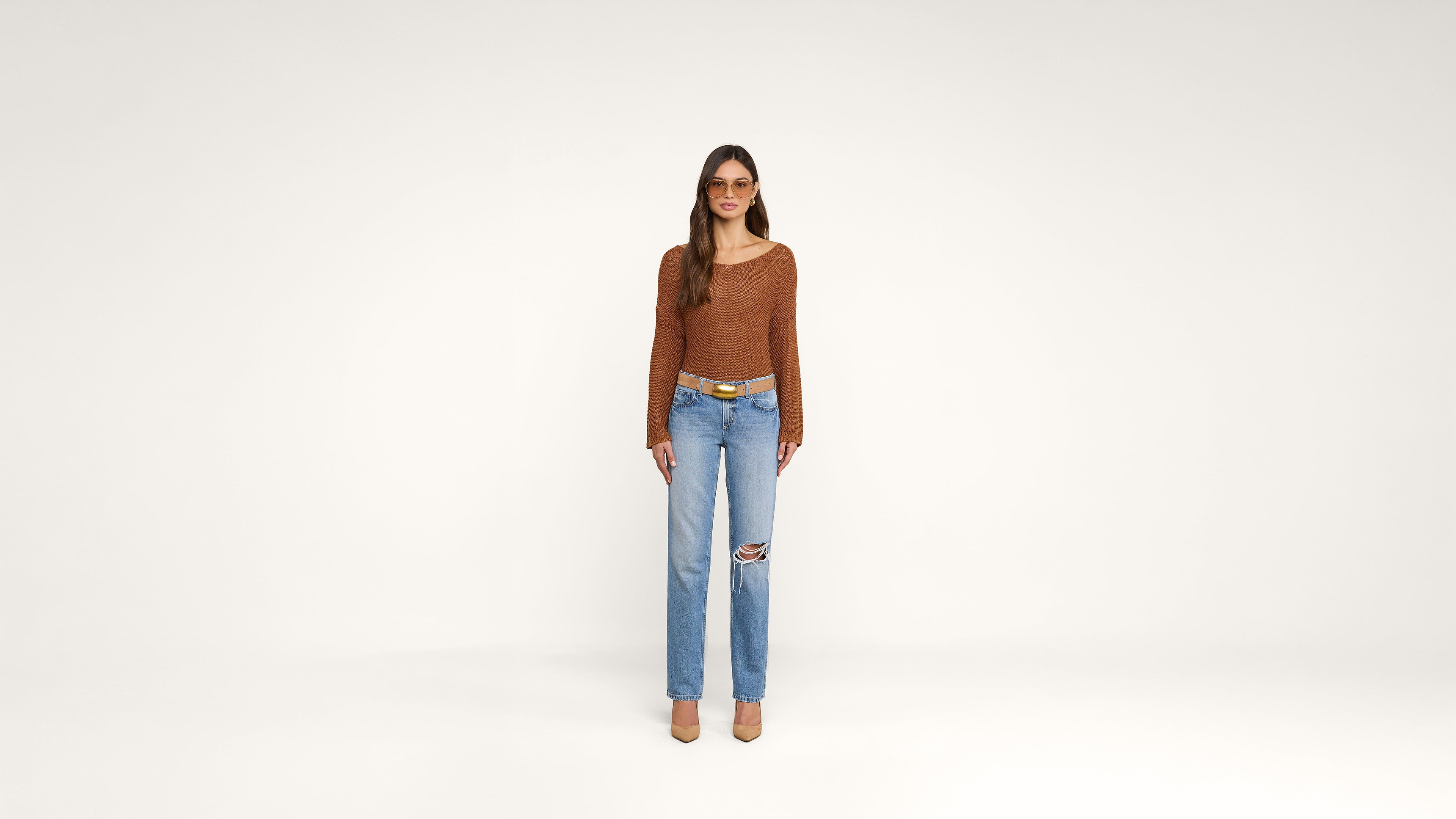 Woman stands facing forward against a plain white background, wearing a brown long-sleeve top, blue ripped jeans, tan heels, a tan belt, and sunglasses. Her hair is straight and loose.