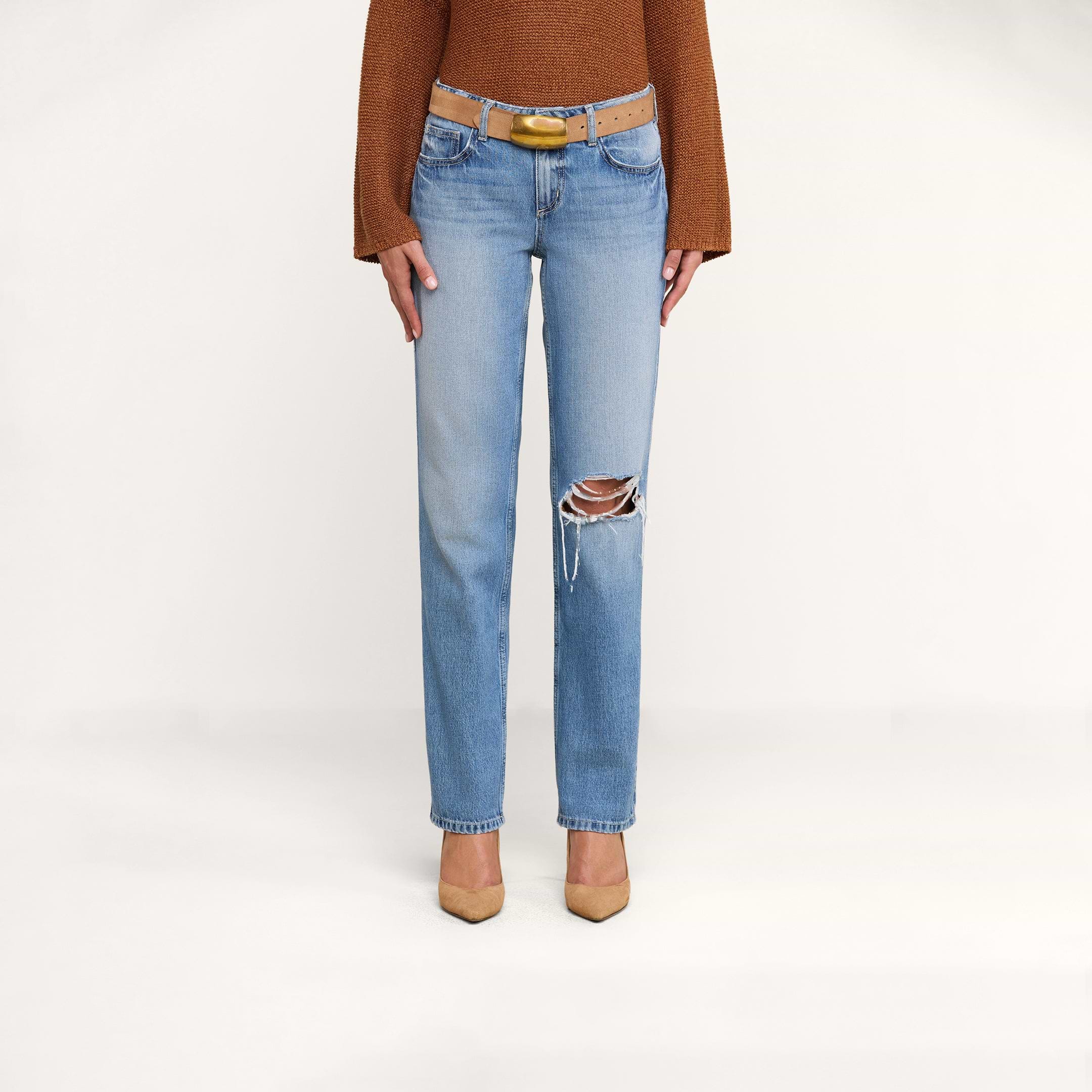 A person wearing a brown long-sleeve top, tan pointed-toe heels, and the Armida Low-Rise Slouch Jean in a vintage indigo wash with a belt, standing against a plain white background.