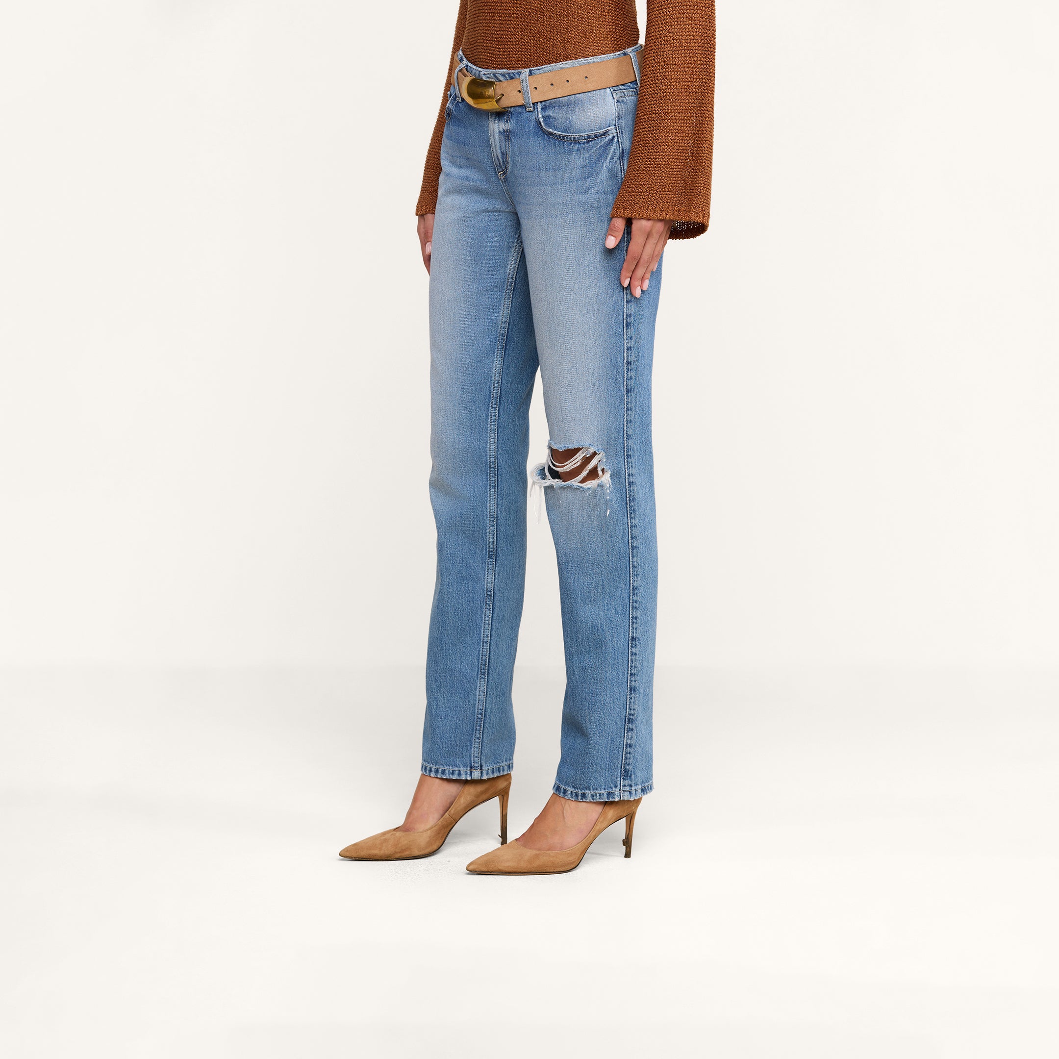 A woman is wearing the Armida Low-Rise Slouch Jean in light blue with a ripped knee, paired with a brown belt, a brown long-sleeve top, and brown pointed-toe heels, standing against a plain white background.
