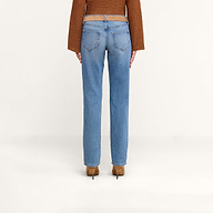 A person is standing with their back to the camera against a plain white background, wearing the Armida Low-Rise Slouch Jean in vintage indigo, paired with a brown sweater and brown heeled shoes.