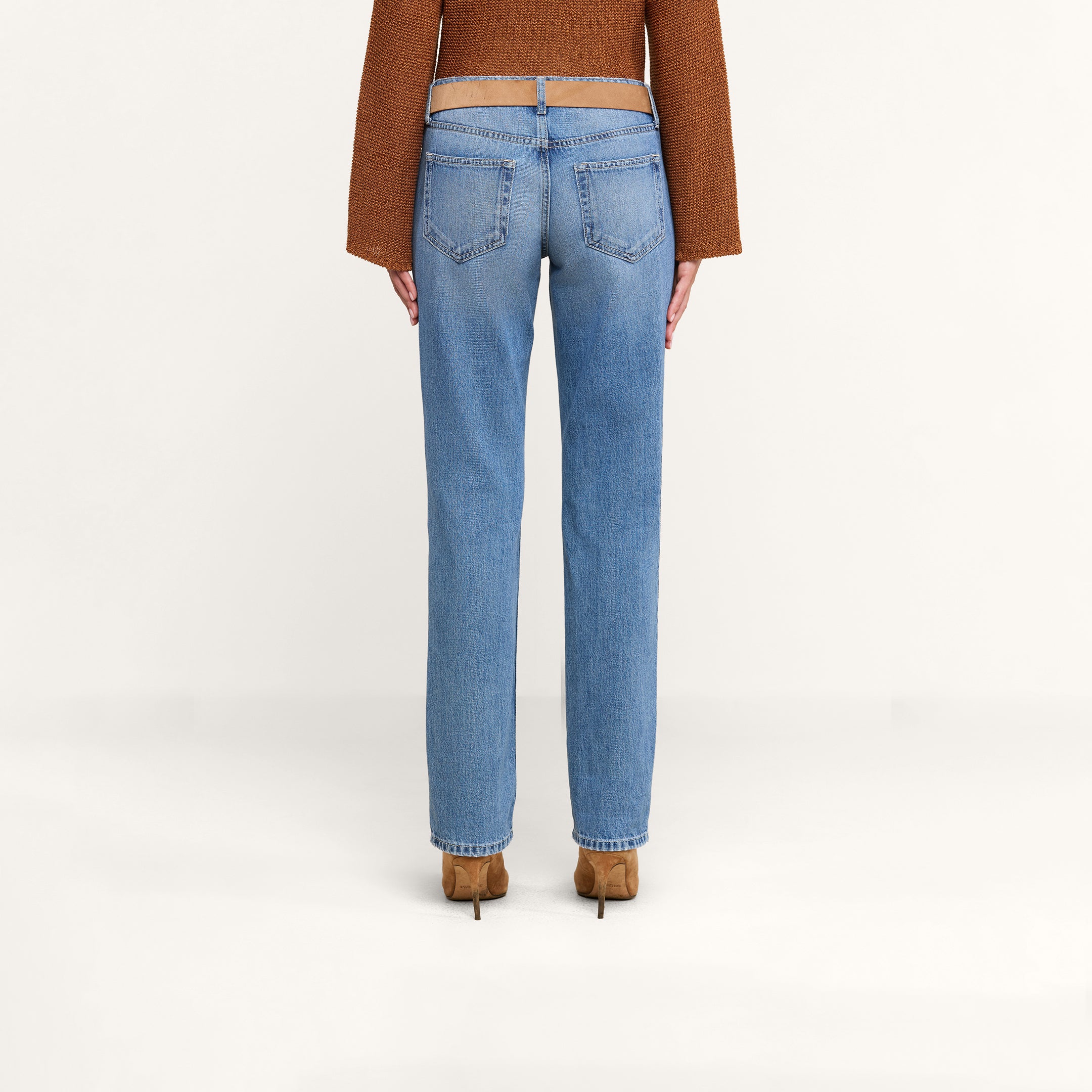 A person is standing with their back to the camera against a plain white background, wearing the Armida Low-Rise Slouch Jean in vintage indigo, paired with a brown sweater and brown heeled shoes.