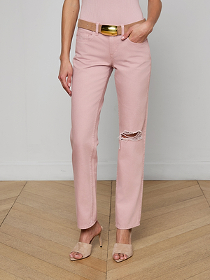 A person stands on a wooden floor wearing the Armida Low-Rise Slouch Jean in light pink with a ripped knee, paired with a beige belt featuring a shiny gold buckle and beige high-heeled sandals. The background is a plain white wall.