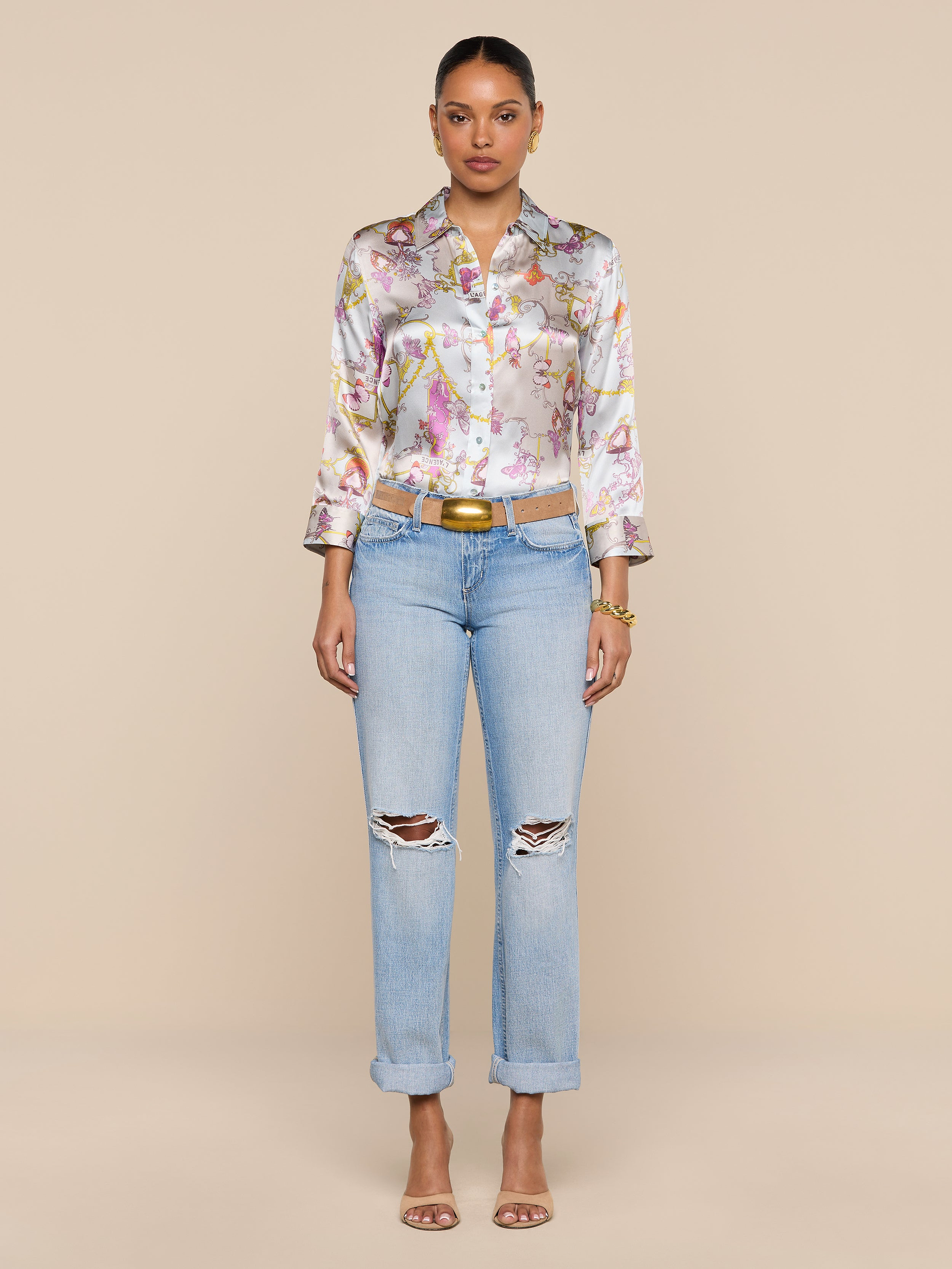 A woman stands facing forward against a plain white background, wearing a vintage-inspired floral button-up shirt, the Ashby Low-Rise Slouch Jean in light blue with ripped details, a belt, and heeled sandals.