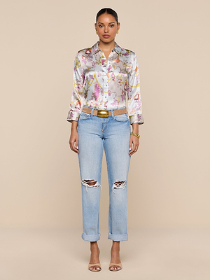 A woman faces forward in a colorful silk blouse, Ashby Low-Rise Slouch Jeans with cuffs, a tan belt, gold jewelry, and beige heeled sandals against a plain beige background.