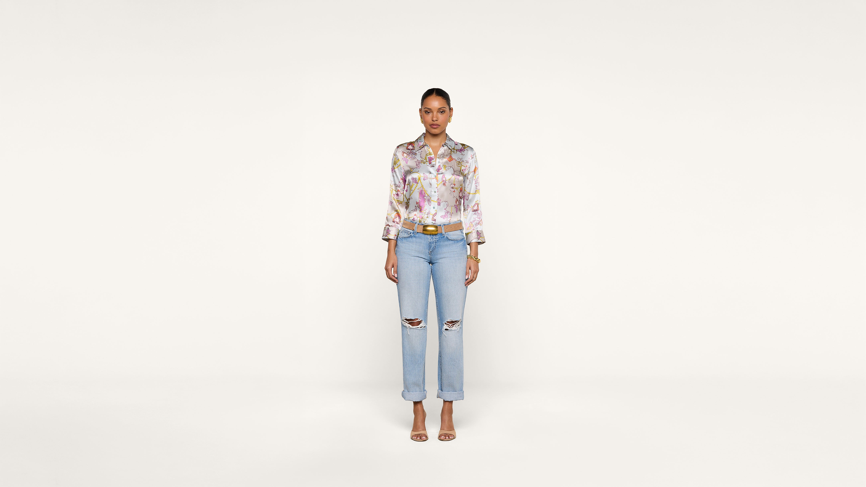 A woman stands facing forward against a plain white background, wearing a floral blouse, ripped light blue jeans, and open-toe heels.