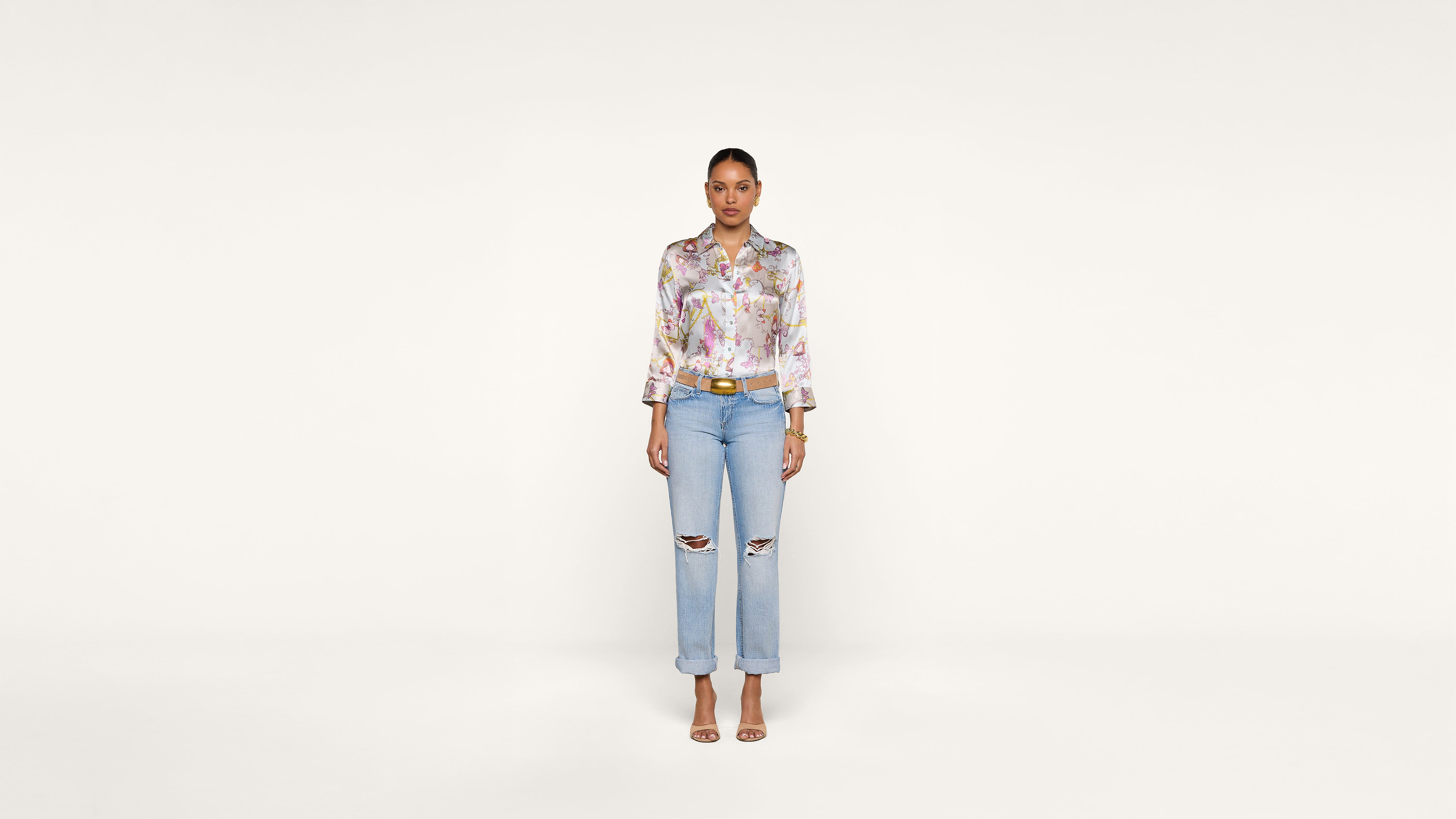 A woman stands facing forward against a plain white background, wearing a floral blouse, ripped light blue jeans, and open-toe heels.