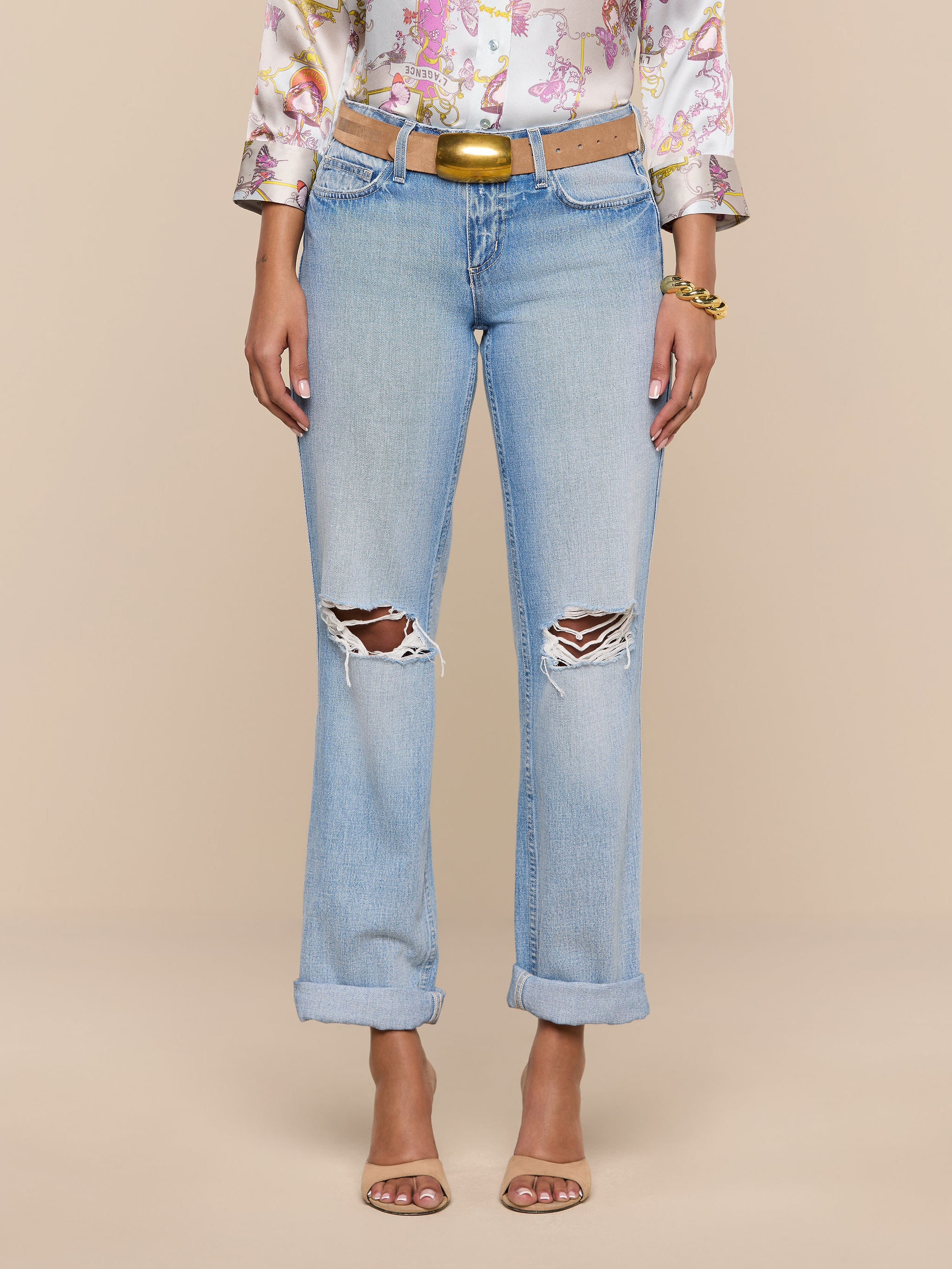 A woman models the Ashby Low-Rise Slouch Jean in light blue, featuring vintage-inspired rips and rolled cuffs. She pairs them with a floral blouse, brown belt, bracelets, and open-toe heels. Her hands are at her sides against a plain white background; her face is not visible.