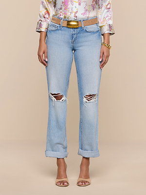 A person wears the Ashby Low-Rise Slouch Jean in light blue with ripped details and rolled cuffs, paired with a brown belt, patterned long-sleeve top, gold bracelet, and open-toed heels, standing against a plain beige background.