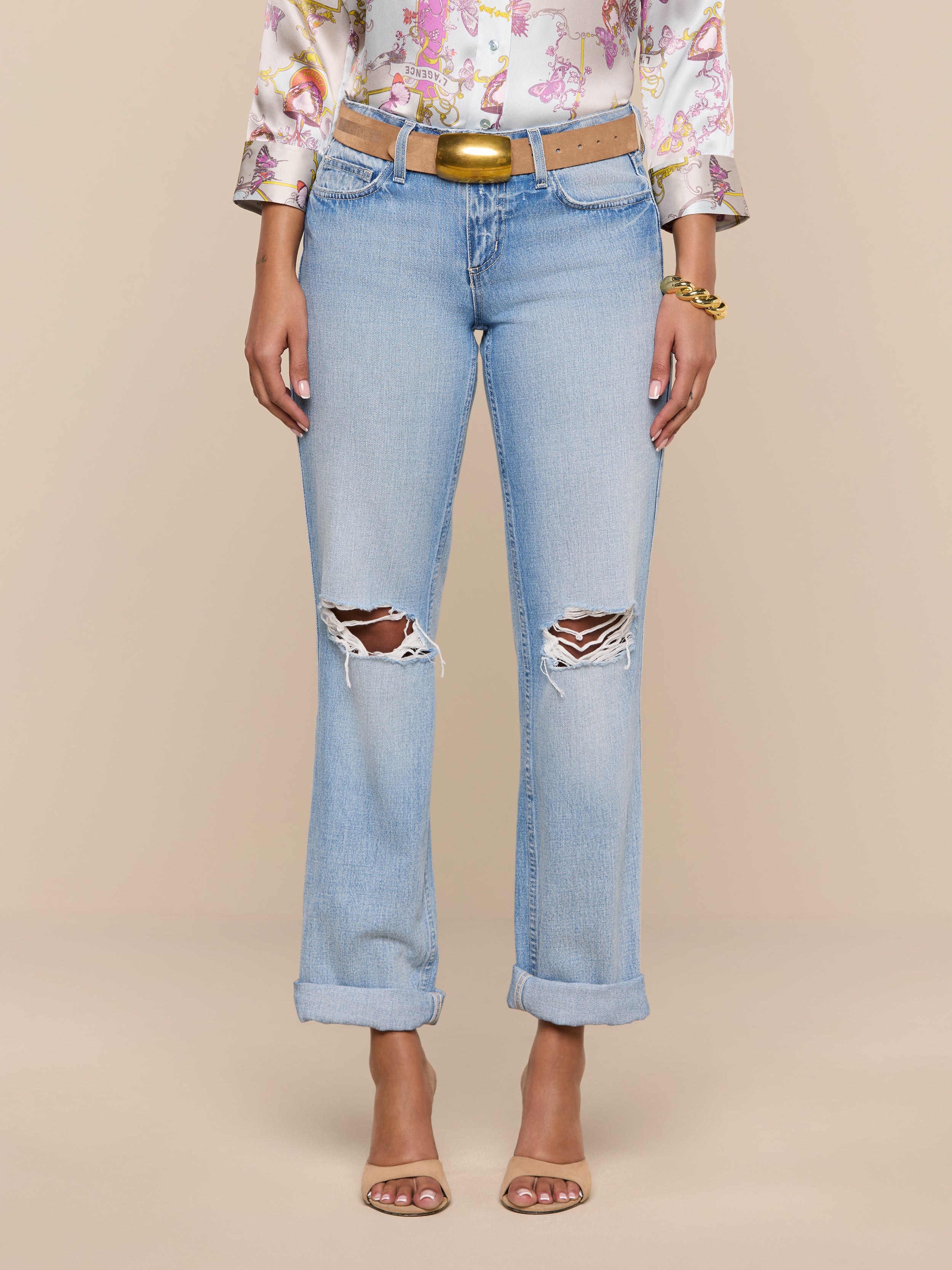A woman models the Ashby Low-Rise Slouch Jean in light blue, featuring vintage-inspired rips and rolled cuffs. She pairs them with a floral blouse, brown belt, bracelets, and open-toe heels. Her hands are at her sides against a plain white background; her face is not visible.