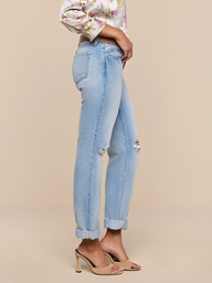 Wearing the Ashby Low-Rise Slouch Jean in vintage-inspired light blue with a small knee rip and cuffed hems, styled with a floral blouse and beige high-heeled sandals, the person stands sideways against a plain white background.