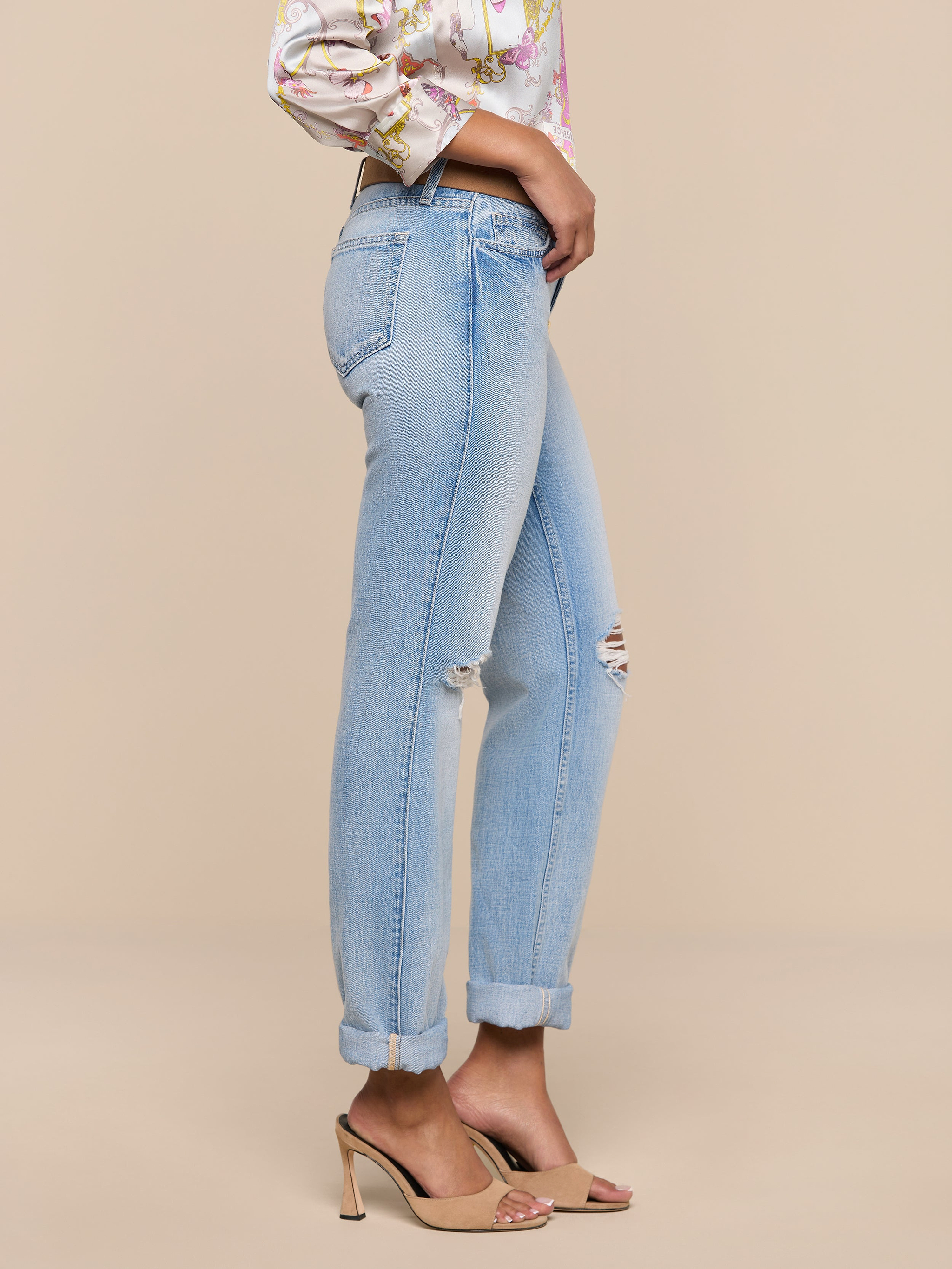 Wearing the Ashby Low-Rise Slouch Jean in vintage-inspired light blue with a small knee rip and cuffed hems, styled with a floral blouse and beige high-heeled sandals, the person stands sideways against a plain white background.