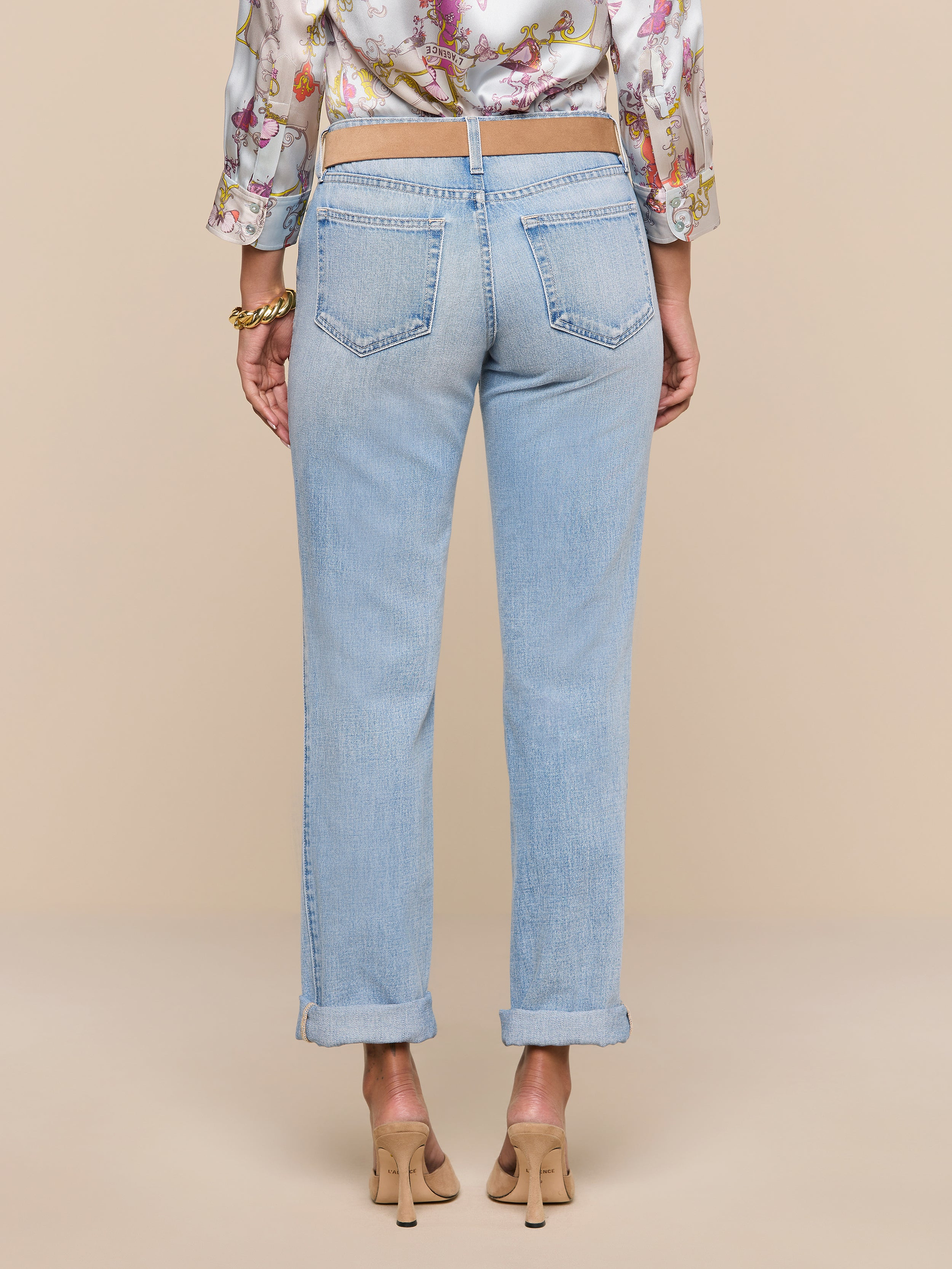 A person stands facing away against a plain white background, wearing the Ashby Low-Rise Slouch Jean in a vintage-inspired light wash, paired with a floral blouse and beige heels.