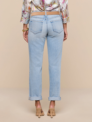 A person stands facing away, wearing the Ashby Low-Rise Slouch Jean in light blue cotton denim, paired with a patterned blouse, beige belt, gold bracelet, and tan high-heeled sandals against a plain beige background.
